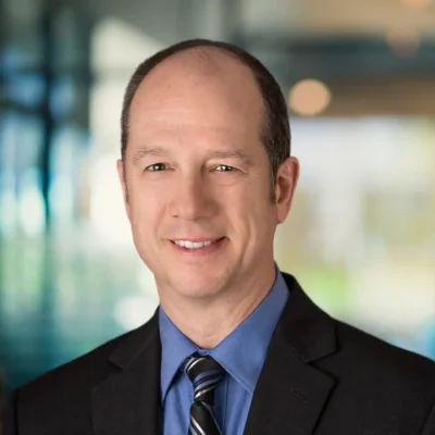 A middle-aged man in a suit and tie smiles at the camera with a blurred office background.