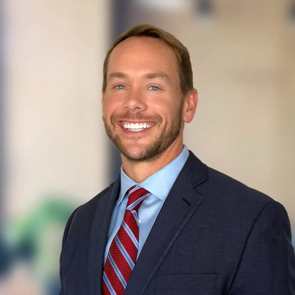 A man in a navy suit, blue shirt, and red striped tie smiles at the camera against a blurred indoor background.