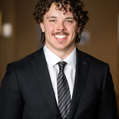 A young man with curly brown hair and a mustache smiles while wearing a black suit, white shirt, and striped tie, standing in front of a blurred brown background.