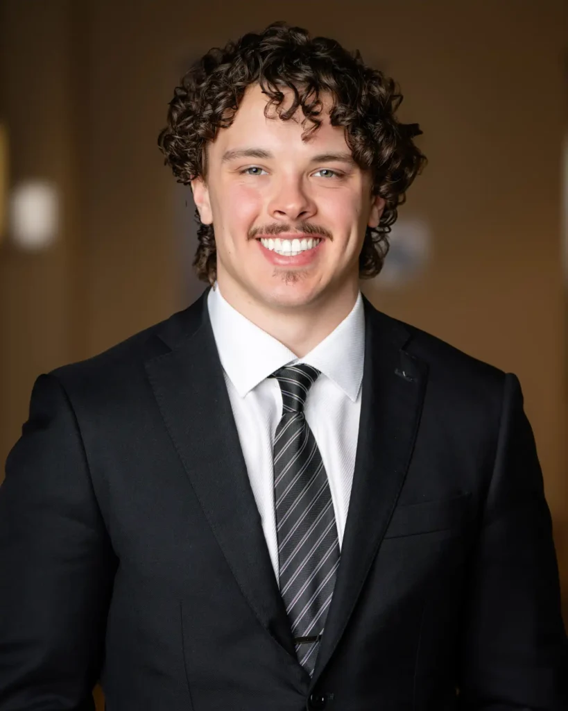 A young man with curly brown hair and a mustache smiles while wearing a black suit, white shirt, and striped tie, standing in front of a blurred brown background.