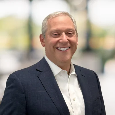 A man in a dark suit and white shirt smiles while standing indoors, with a blurred background of windows and greenery.