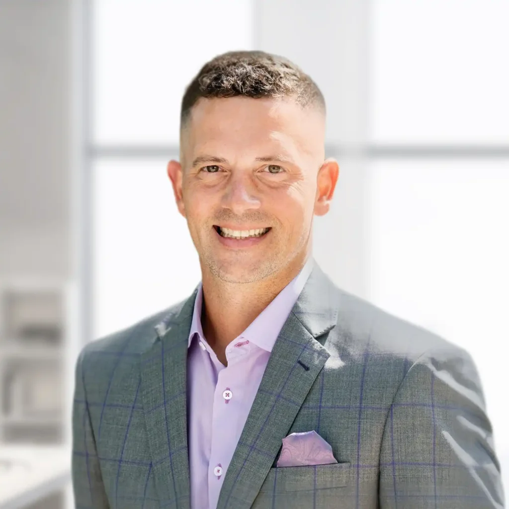 A man in a light gray checked suit and lavender shirt stands smiling in a bright, modern office setting.