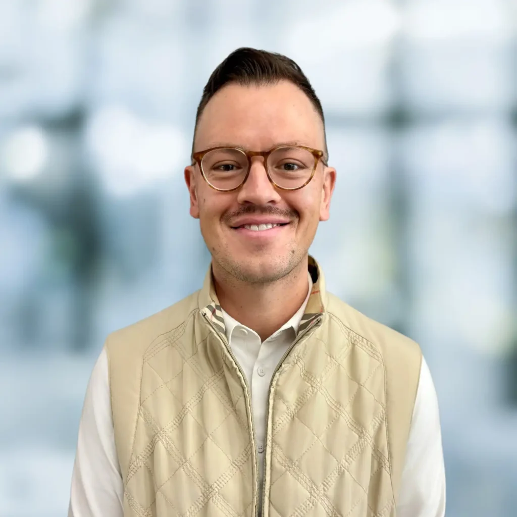 A man with short hair, glasses, and a mustache smiles while wearing a beige quilted vest over a white shirt, standing in front of a blurred background.