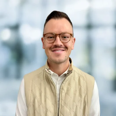 A man with short hair, glasses, and a mustache smiles while wearing a beige quilted vest over a white shirt, standing in front of a blurred background.