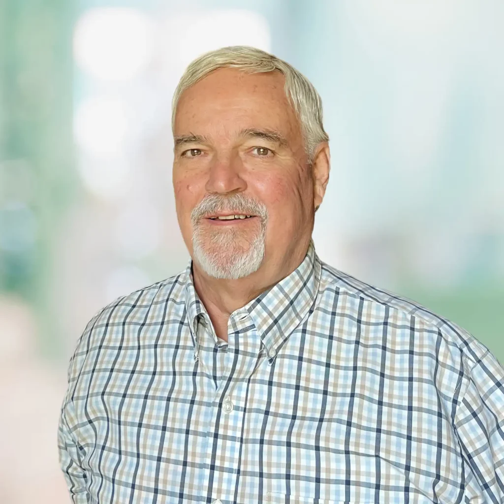 An older man with gray hair and a beard is wearing a blue and white plaid shirt, standing in front of a blurred background.