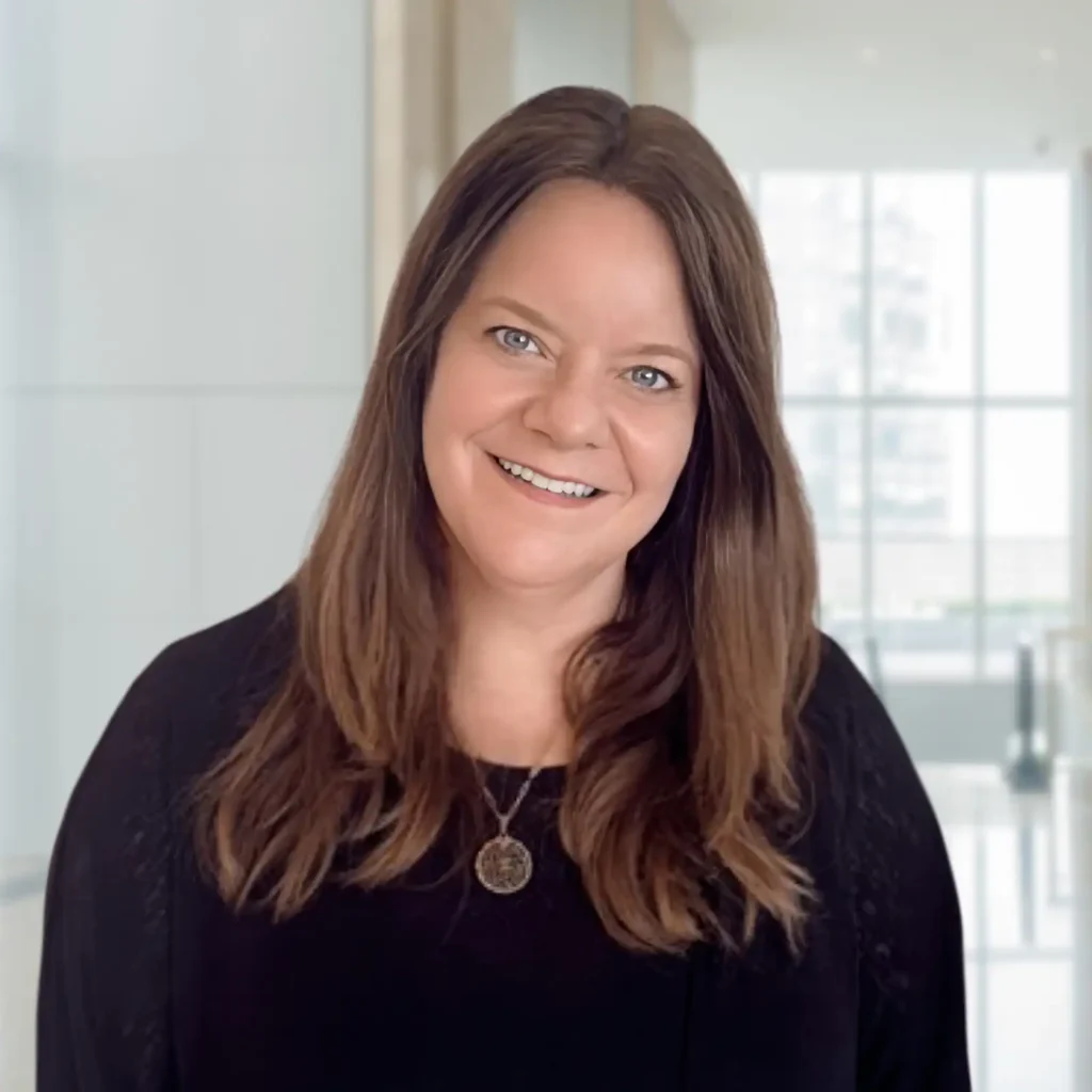 A woman with long brown hair, wearing a black top and a pendant necklace, smiles while standing in a bright, modern indoor setting with large windows in the background.