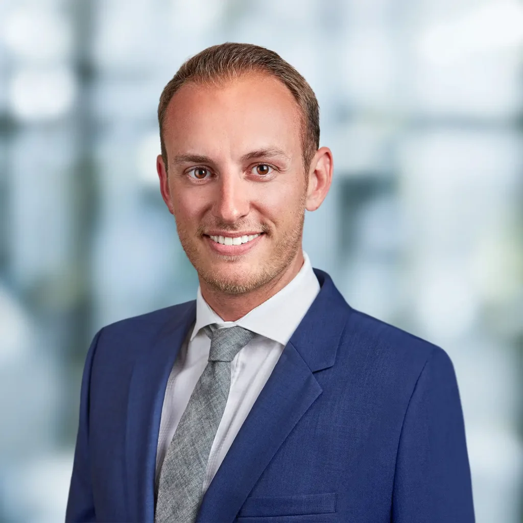 A man in a blue suit and gray tie smiles at the camera with a blurred office background.