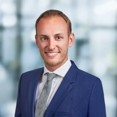 A man in a blue suit and gray tie smiles at the camera with a blurred office background.