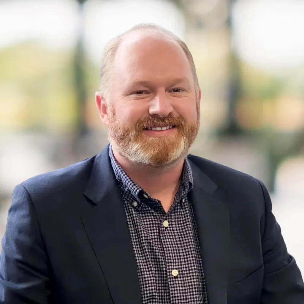 A man with a reddish beard and hair, wearing a dark blazer and checkered shirt, smiles at the camera with a blurred background.