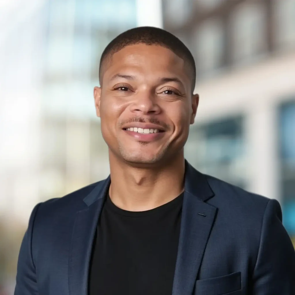 A man wearing a dark blazer and black shirt smiles at the camera, standing outdoors with a blurred cityscape in the background.