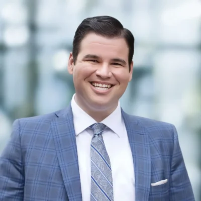 A man in a blue plaid suit with a patterned tie and white shirt is smiling at the camera, standing in front of a blurred background.