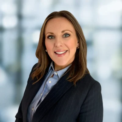 A woman with shoulder-length brown hair wearing a dark pinstripe blazer and blue shirt smiles at the camera against a blurred background.