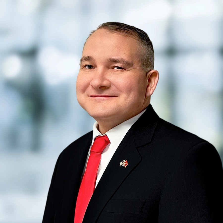 A man in a black suit, white shirt, and red tie is smiling at the camera against a blurred light background.