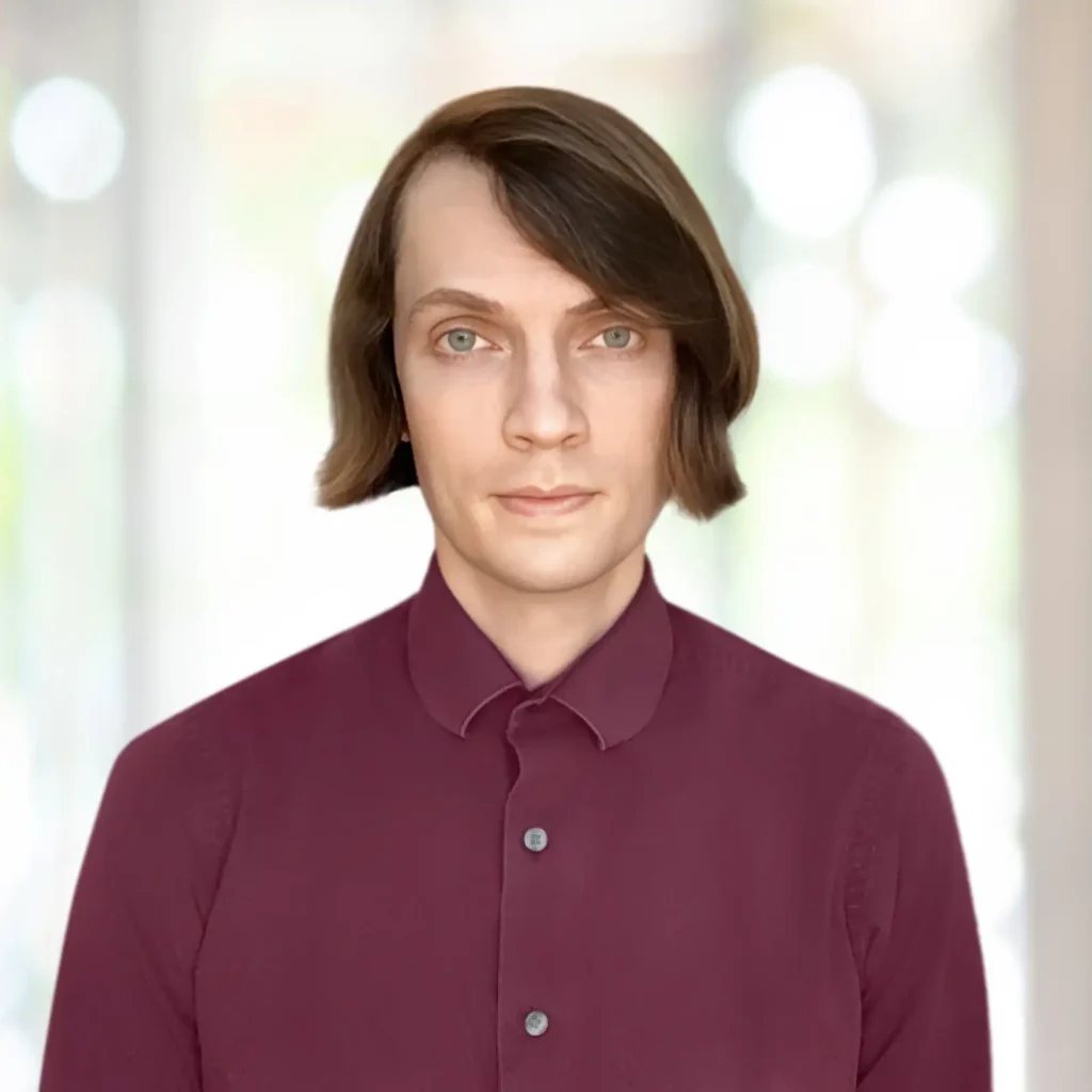 A person with straight brown hair, fair skin, and light-colored eyes is wearing a maroon button-up shirt, standing in front of a blurred light background.