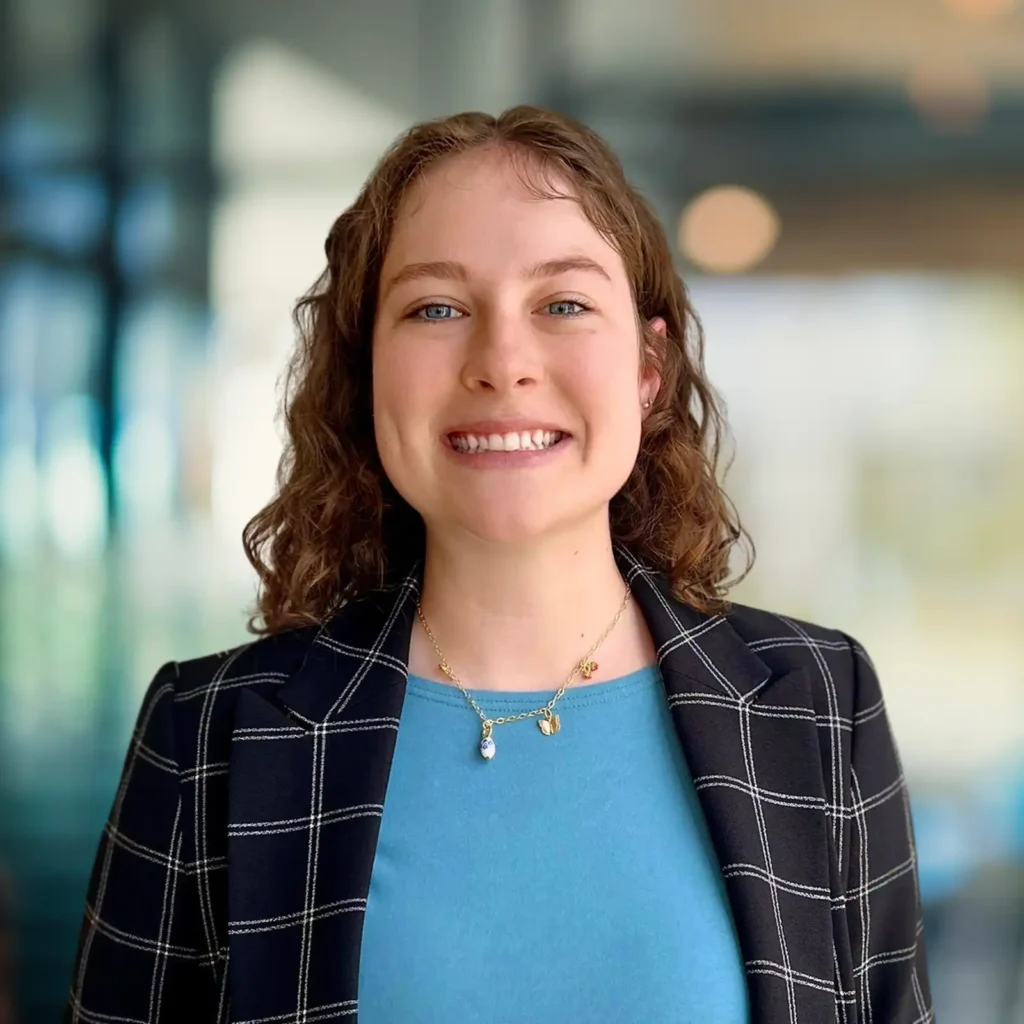 A woman with curly brown hair, wearing a blue shirt and plaid blazer, smiles at the camera in a brightly lit indoor setting.