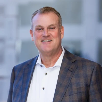 A middle-aged man in a checked blazer and white shirt smiles while standing indoors with a blurred background.