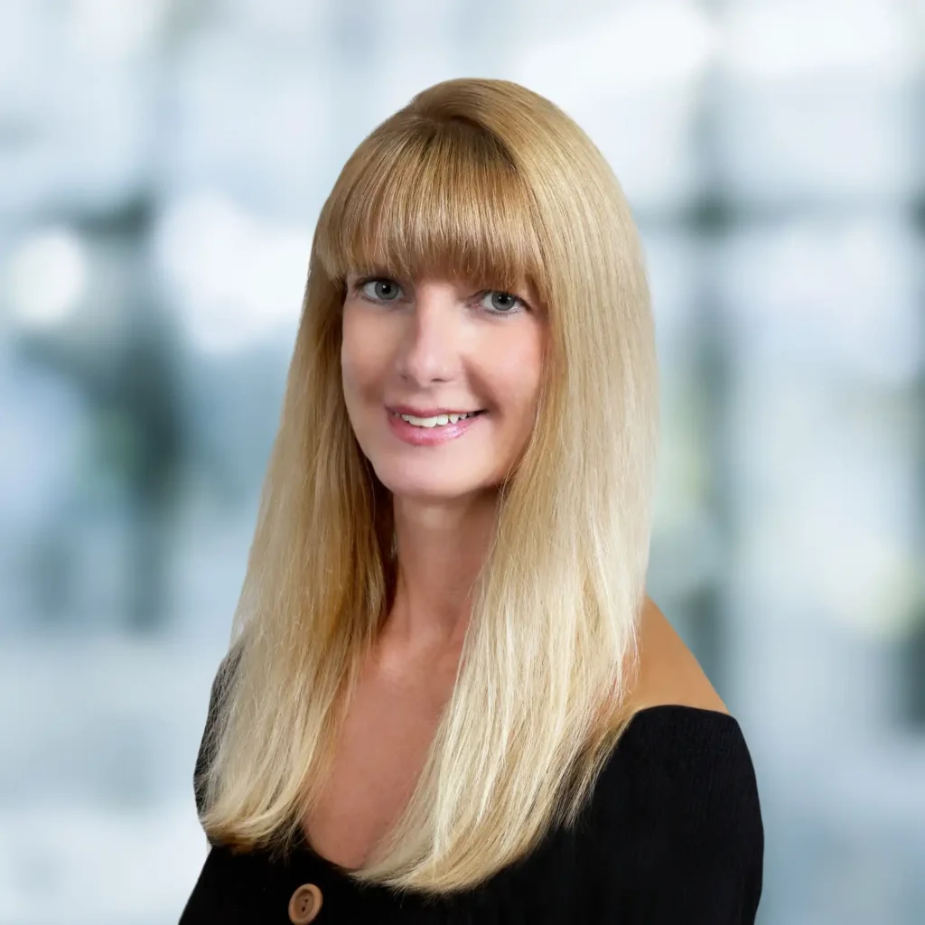 A woman with straight, long blonde hair and bangs is smiling at the camera. She is wearing a black off-the-shoulder top and is posed against a blurred light background.