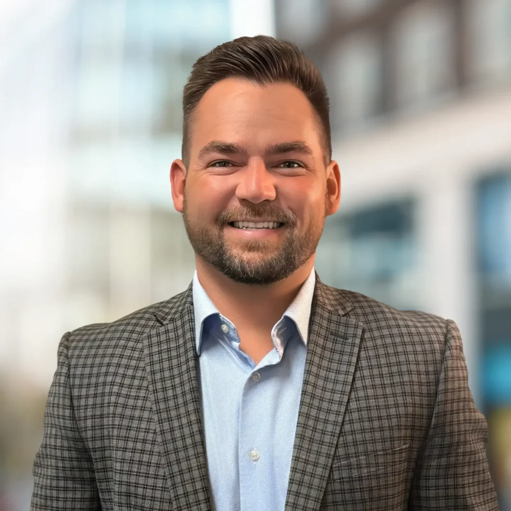 A man with short brown hair and a beard, wearing a checked suit jacket over a light blue shirt, stands smiling in front of a blurred urban background.