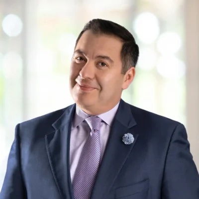 A man wearing a dark blue suit, light purple shirt, patterned tie, and a flower lapel pin stands indoors with a blurred background.