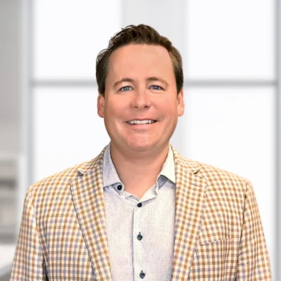 A man with short brown hair, wearing a light plaid blazer and light-colored shirt, smiles in front of a blurred office background.