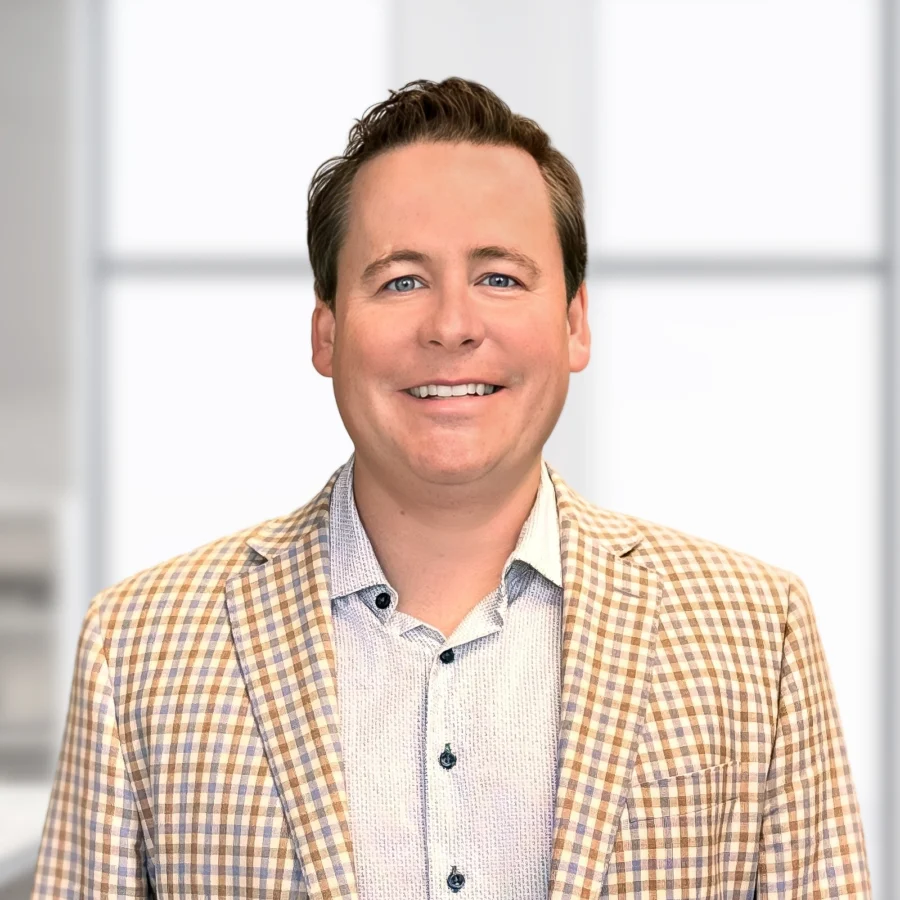 Man wearing a light plaid blazer and collared shirt smiles while standing in a brightly lit, modern office setting.