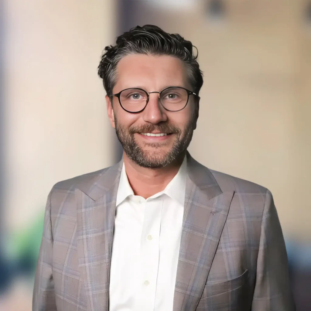 A man with short, wavy hair and a beard wearing glasses, a light plaid suit jacket, and a white shirt, standing against a blurred background.
