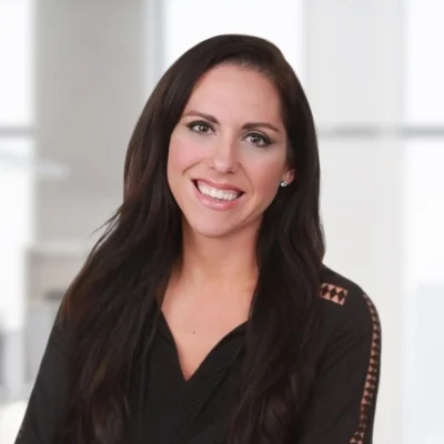 Woman with long dark hair, wearing a black top, smiling, posed in a brightly lit, modern office setting.
