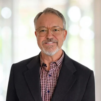 A middle-aged man with gray hair, glasses, and a beard is wearing a dark blazer over a checkered shirt, standing in front of a blurred light background.