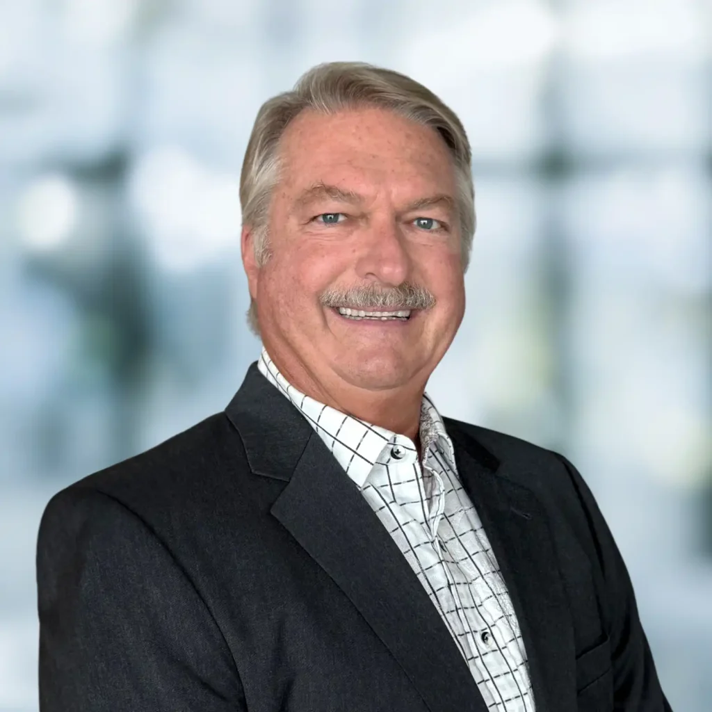 A man with gray hair and a mustache wearing a dark suit jacket and a white checkered shirt, smiling at the camera against a blurred background.