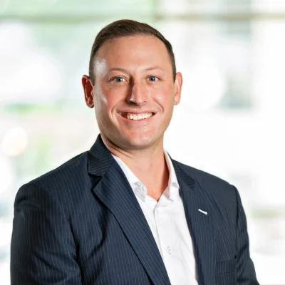 A man in a pinstripe suit jacket and white shirt smiling, posing for a professional portrait against a blurred light background.