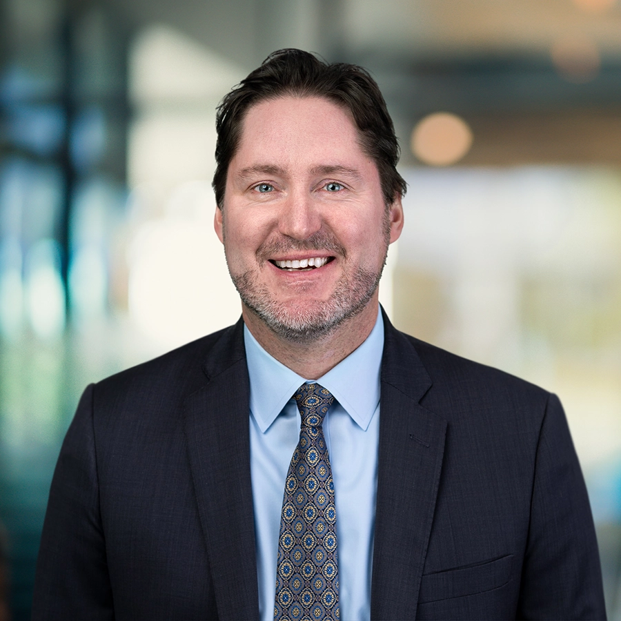 A man in a suit and tie is smiling, standing in front of a blurred office background.