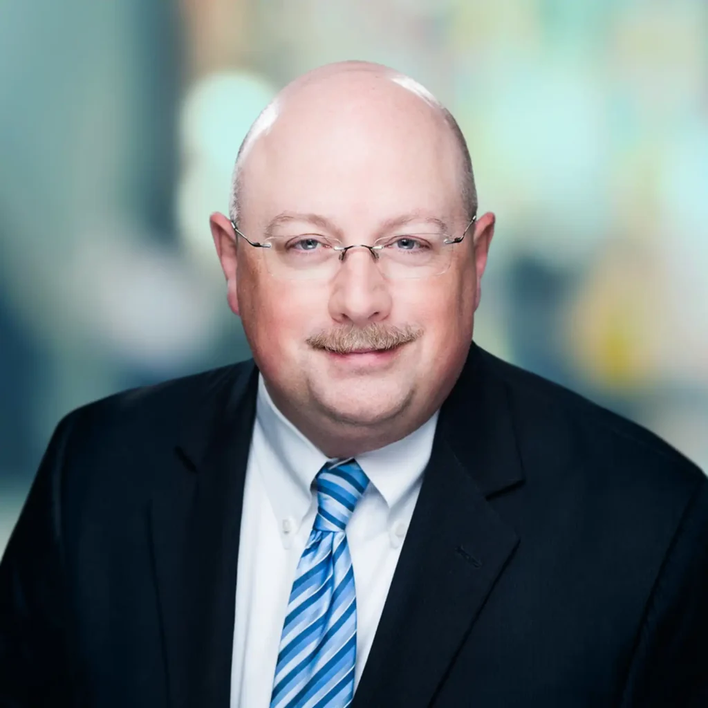 A middle-aged man with glasses, a mustache, and a bald head is wearing a dark suit, white shirt, and blue striped tie, posed against a blurred background.