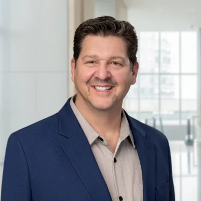 A man with short brown hair and a mustache, wearing a blue blazer and light striped shirt, smiles in a modern office setting with large windows in the background.
