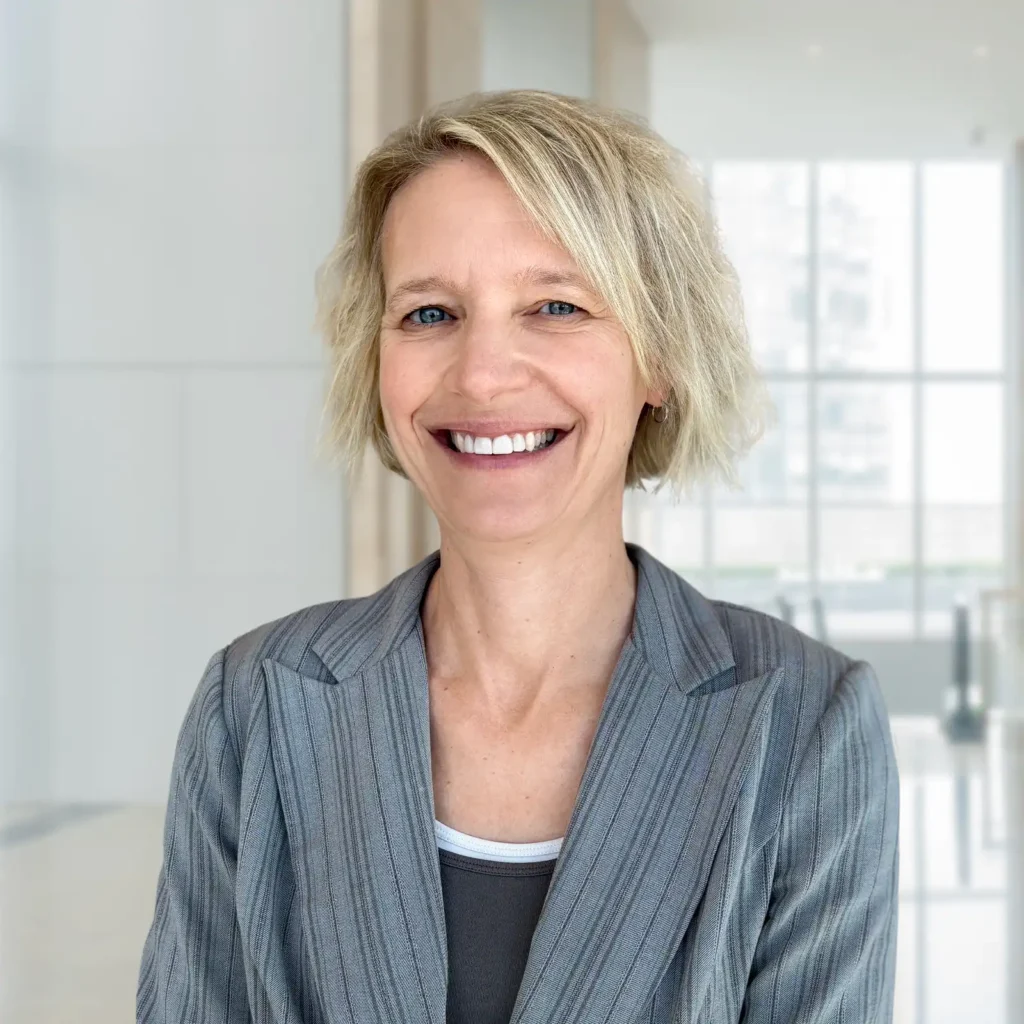 A woman with short blonde hair in a gray pinstripe blazer smiles while standing in a bright, modern office space.