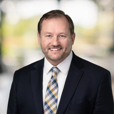 A middle-aged man with short brown hair and a trimmed beard, wearing a dark suit, white shirt, and patterned tie, standing in front of a blurred background.