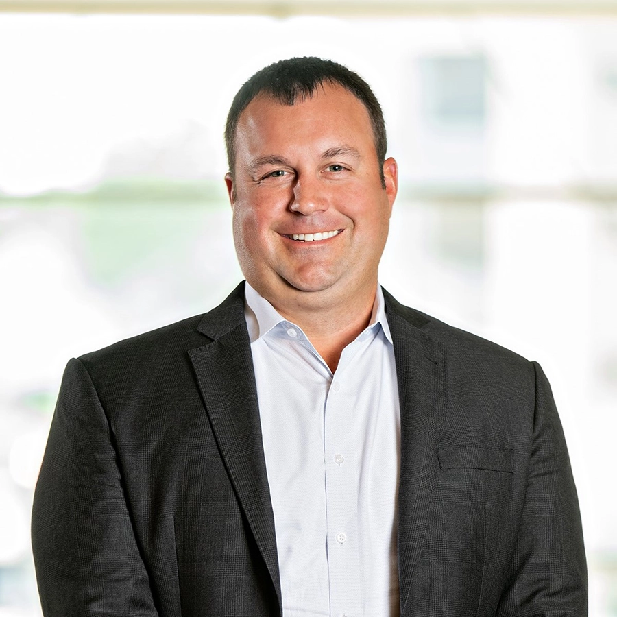 A man in a dark suit jacket and white shirt stands smiling in front of a bright, blurred background.