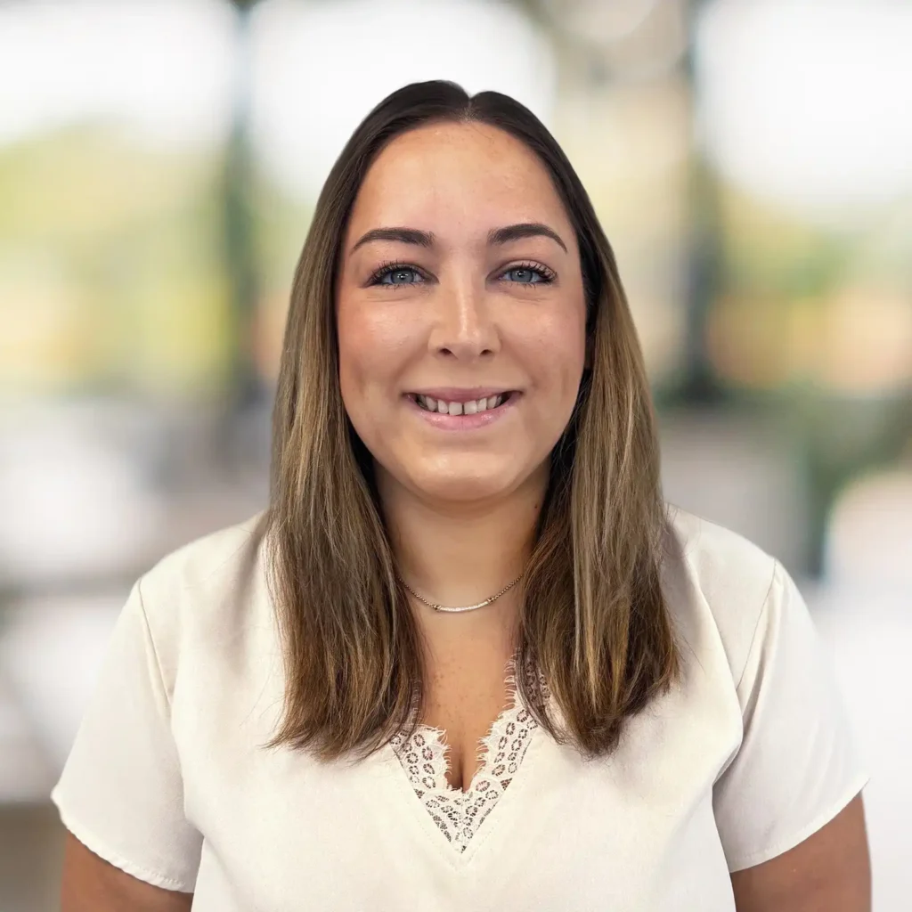 A woman with straight brown hair and a light blouse smiles at the camera, standing in front of a blurred background.