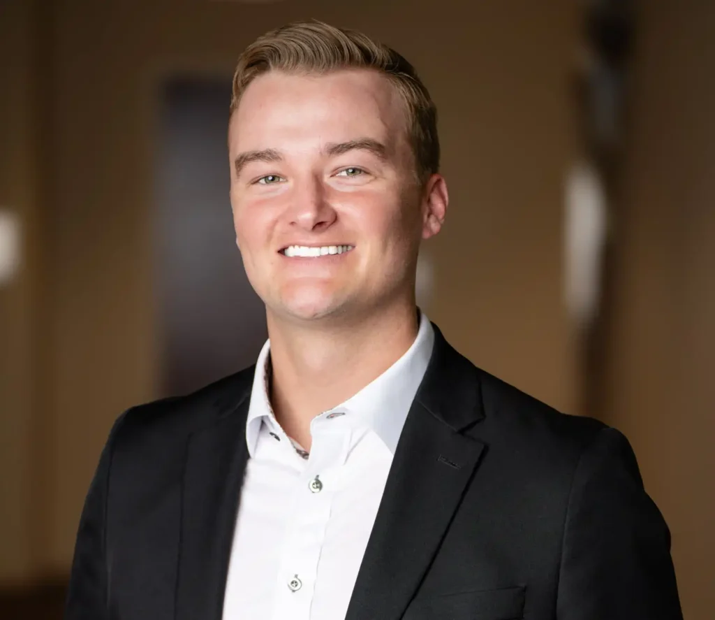 A man wearing a black suit jacket and white collared shirt stands smiling in front of a blurred indoor background.