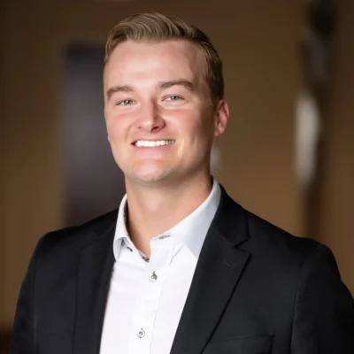 A man wearing a black suit jacket and white collared shirt stands smiling in front of a blurred indoor background.