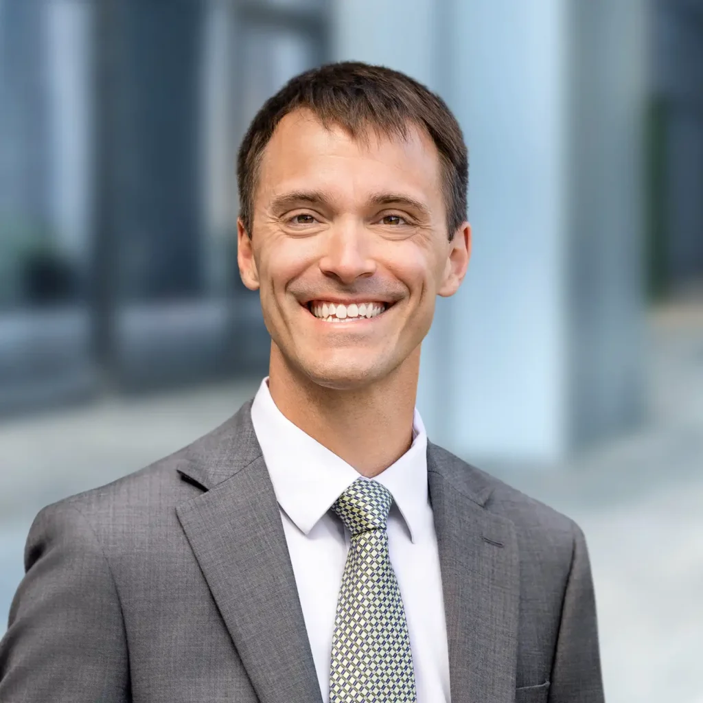 A man in a gray suit and patterned tie smiles while standing outdoors in front of a blurred office building background.
