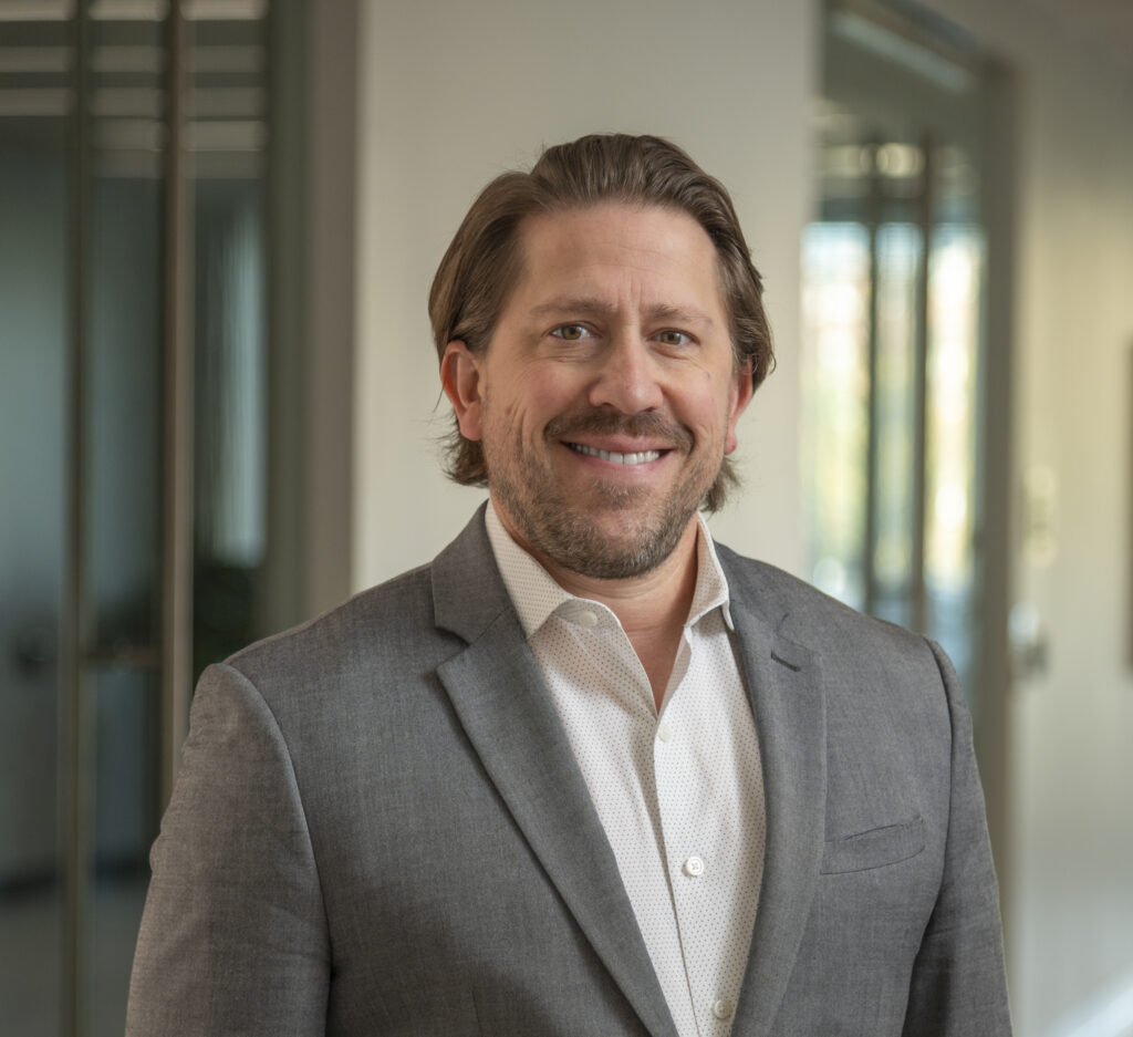 A man in a gray suit and white shirt stands in a well-lit hallway, smiling at the camera.