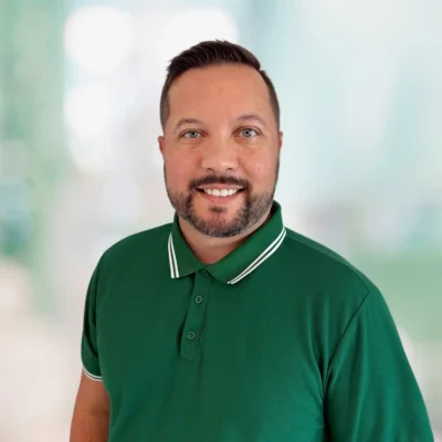 A man with short brown hair and a beard, wearing a green collared shirt, smiles at the camera against a blurred light background.