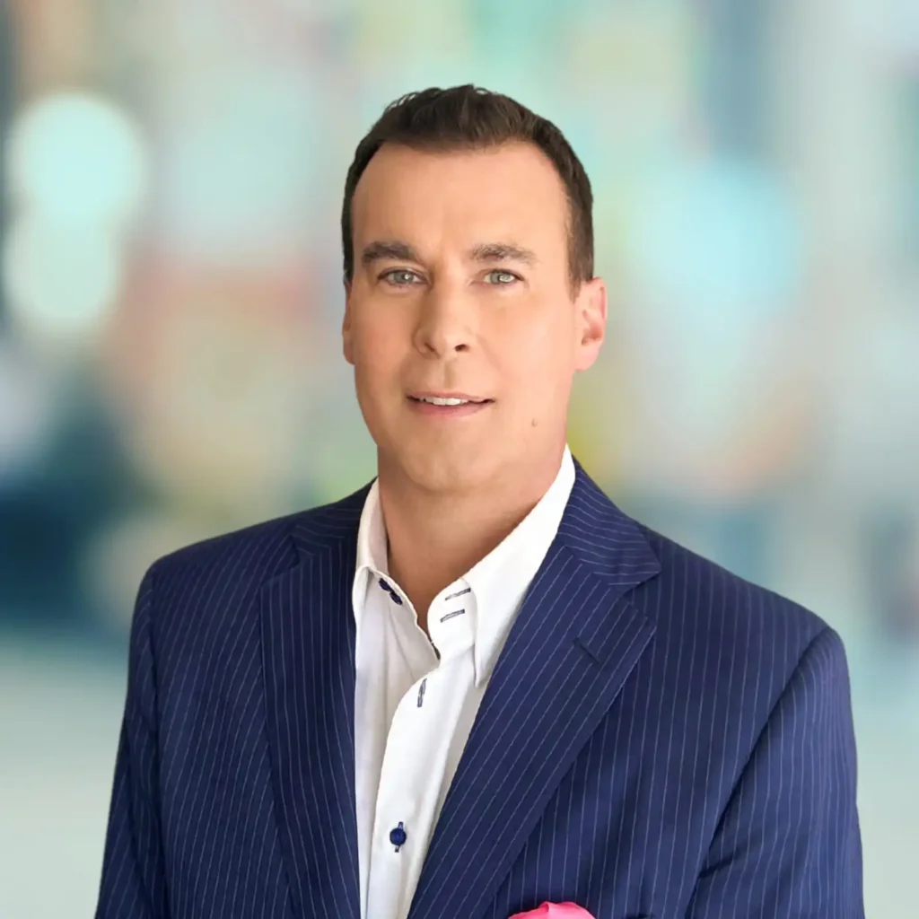 A man wearing a blue pinstripe suit and white shirt stands in front of a blurred, colorful background.