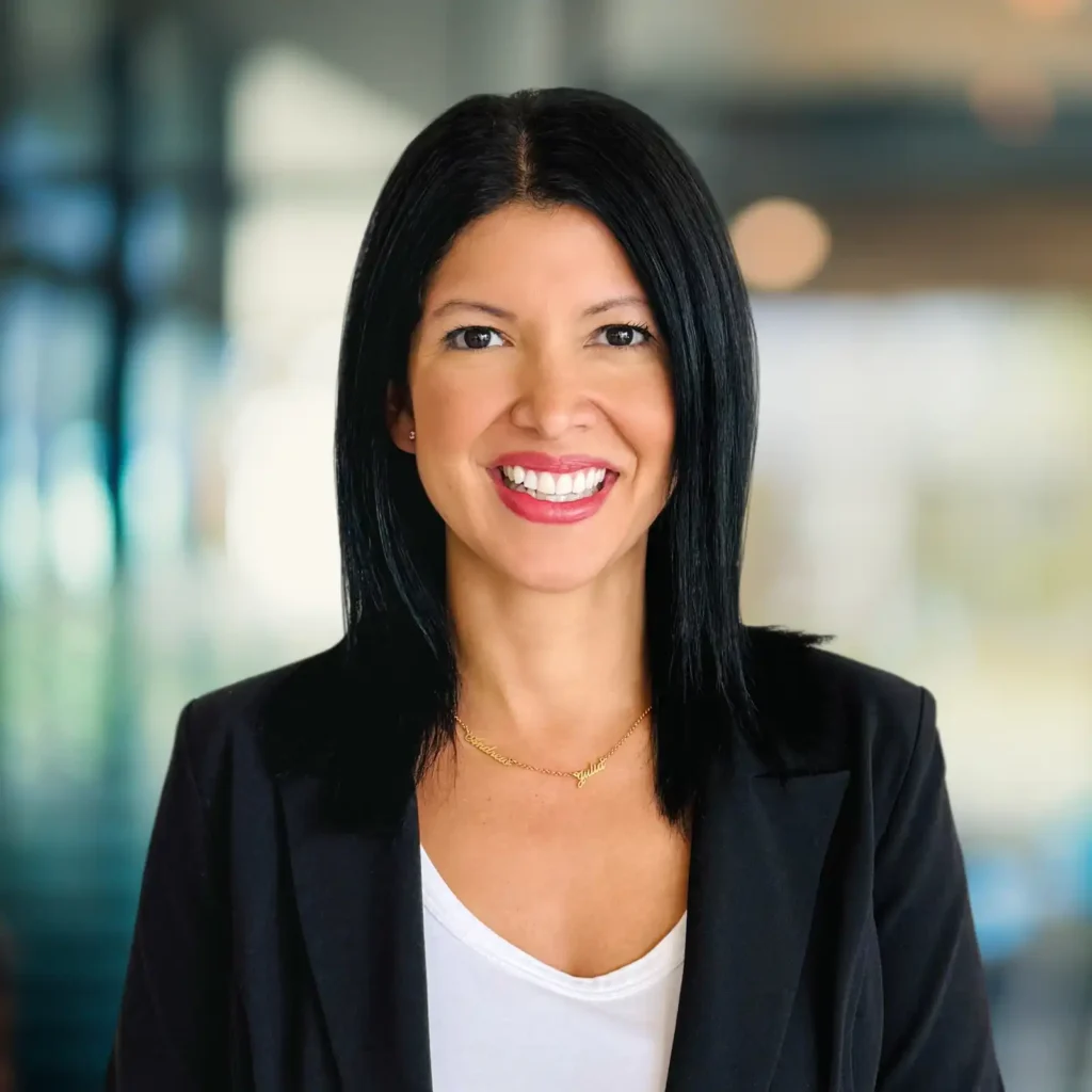A woman with straight black hair, wearing a black blazer over a white top, smiles at the camera against a blurred indoor background.