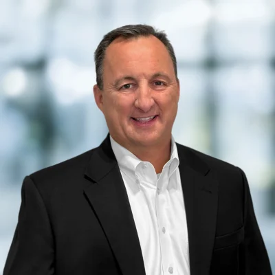 Man in a black suit jacket and white dress shirt smiles at the camera, standing in front of a blurred indoor background.