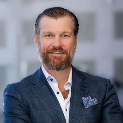 A man with a beard and slicked-back hair, wearing a blue checked suit jacket, white shirt, and patterned pocket square, stands with arms crossed against a blurred background.