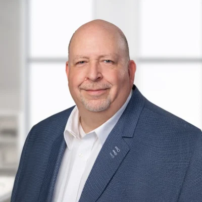 A middle-aged man with a bald head and goatee, wearing a blue blazer and white shirt, poses and smiles in front of a bright, blurred indoor background.
