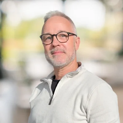 A middle-aged man with short gray hair, a beard, and glasses, wearing a light-colored zip-up sweater, stands in a softly blurred outdoor setting.