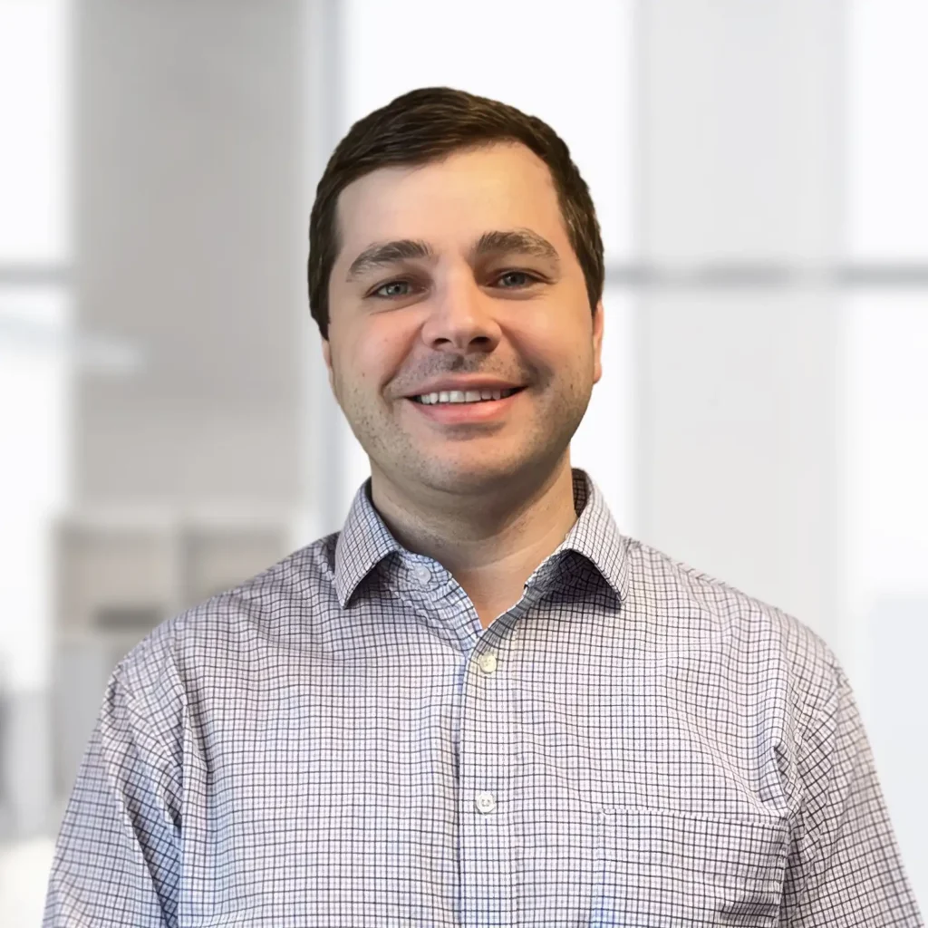 A man with short brown hair, wearing a light checkered shirt, smiles while standing in front of a blurred office background.