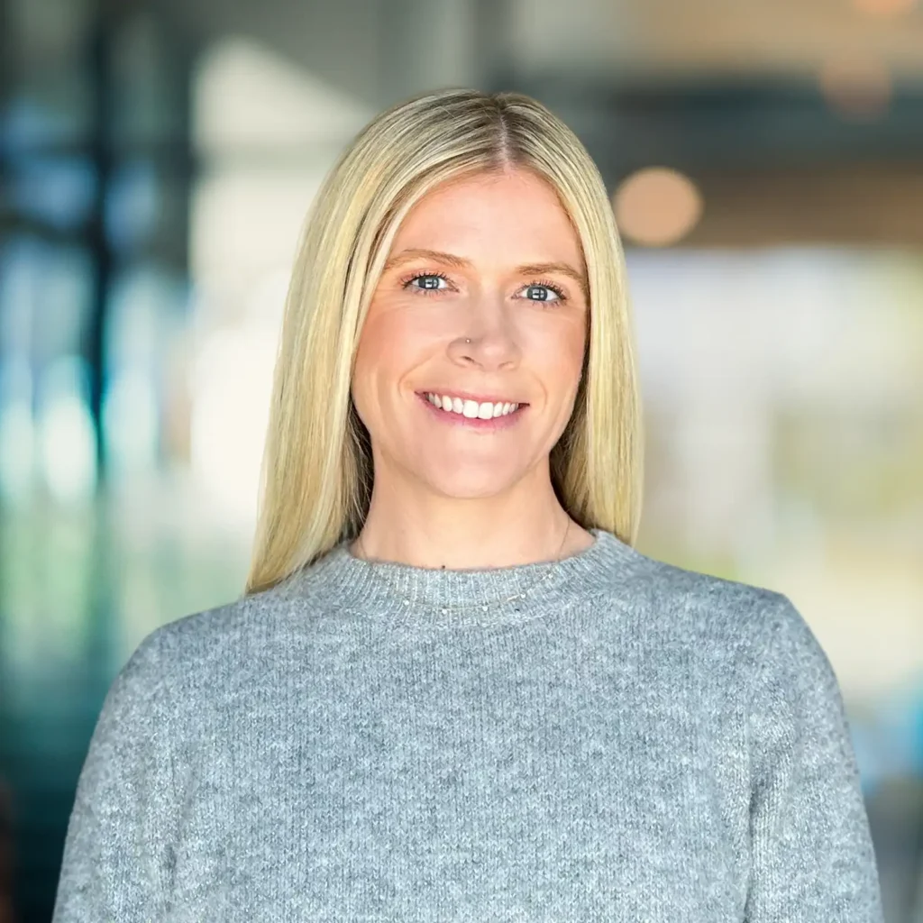 A woman with straight blonde hair and a grey sweater smiles at the camera, standing in a brightly lit indoor setting with a blurred background.